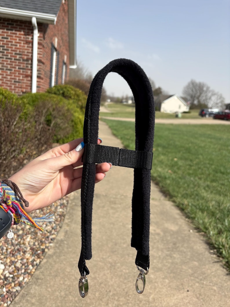 May include: Black shoulder strap with a soft, fuzzy pad and adjustable webbing. The strap has two silver-coloured swivel clips for attachment. The background shows a pavement, greenery, and a house.