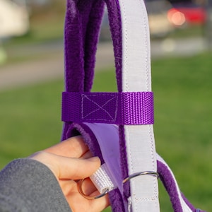 May include: Close-up of a purple and white equestrian halter. The halter features a plush purple lining, a white padded strap, and a purple nylon strap. Silver-coloured metal hardware is visible, including a snap hook and a ring.