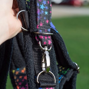 May include: Black dog harness with a vibrant star pattern on the straps. The straps feature silver metal hardware and a secure clasp. The harness is made from a soft, padded material for comfort.