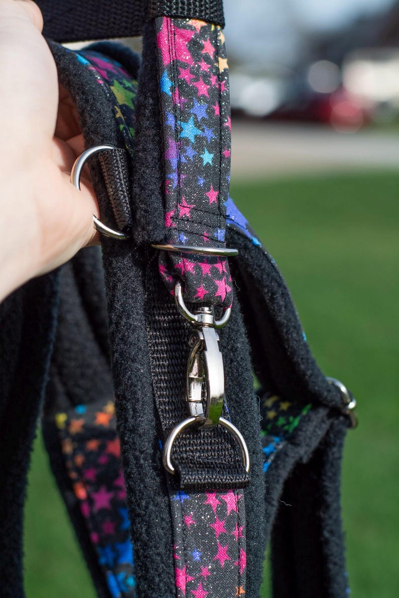 May include: Black dog harness with a vibrant star pattern on the straps. The straps feature silver metal hardware and a secure clasp. The harness is made from a soft, padded material for comfort.