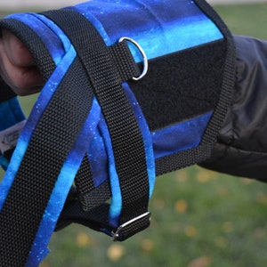May include: A dog harness with a blue and black galaxy print. The harness has black nylon straps, metal hardware, and a black Velcro closure. The harness is designed to fit around the dog's chest and shoulders.