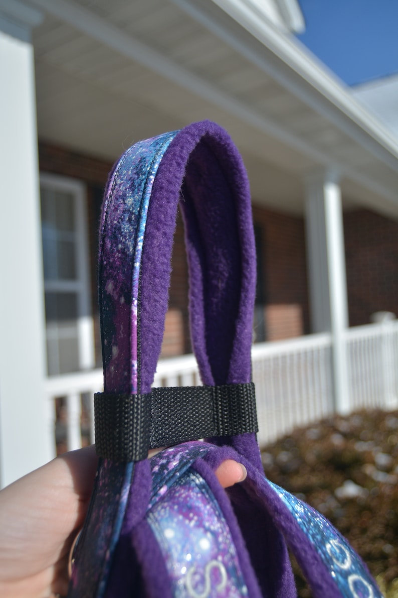May include: A dog harness with a purple and galaxy design, featuring a black nylon strap. The harness has a soft, plush purple lining for comfort. The outer fabric showcases a vibrant design of blues, purples, and white, reminiscent of a starry night.