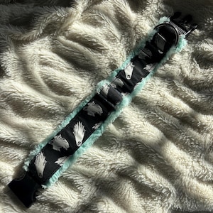 Pode incluir: Uma coleira preta para cães com um padrão de penas brancas. A coleira tem um forro de pelúcia azul claro e uma fivela de plástico preta. Está sobre uma superfície branca e fofa.