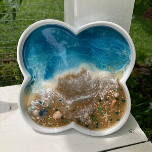 May include: A white ceramic tray shaped like a four-leaf clover with a blue and white resin ocean scene inside. The resin includes white waves, brown sand, and small seashells.