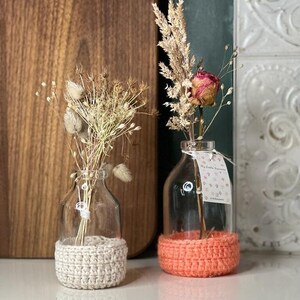 Peut inclure: Deux bouteilles en verre transparent avec des couvercles en crochet. La bouteille de gauche a un couvercle beige et la bouteille de droite a un couvercle corail. Les deux bouteilles sont remplies de fleurs séchées.