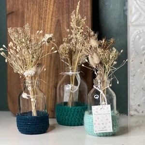 Peut inclure: Trois bouteilles en verre avec des fleurs séchées et des couvercles en crochet. Les couvercles sont turquoise, vert et bleu clair. Le couvercle bleu clair porte une étiquette qui dit "Mes petits trésors".