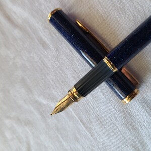 Puede incluir: Un bol&iacute;grafo de fuente azul y dorado con un cuerpo azul texturizado y detalles dorados. El bol&iacute;grafo est&aacute; cerrado y sobre una superficie blanca.