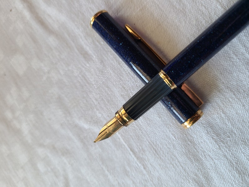 Puede incluir: Un bol&iacute;grafo de fuente azul y dorado con un cuerpo azul texturizado y detalles dorados. El bol&iacute;grafo est&aacute; cerrado y sobre una superficie blanca.