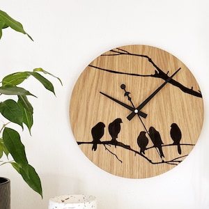Op de afbeelding: Een ronde houten klok met een zwart silhouet van vogels op een tak. De klok heeft zwarte wijzers en een witte achtergrond.
