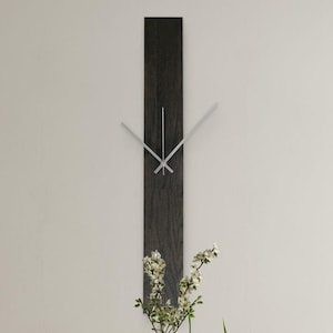 May include: A contemporary wall clock with a dark wood grain effect. The clock has silver hands and a simple design. It's mounted on a light wall, above a table with flowers. The clock's dimensions are approximately 120 cm in height.