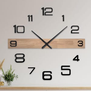 Op de afbeelding: Een moderne wandklok met grote, zwarte cijfers en wijzers. De wijzerplaat van de klok bestaat uit afzonderlijke cijfers en wijzers, met een houten plank in het midden. De klok is gemonteerd op een witte muur boven een witte kast.