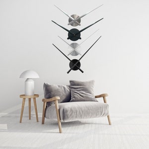 May include: Three sets of clock hands in black, silver, and gold, displayed against a white wall. The hands are arranged vertically, with the black hands at the bottom and the gold hands at the top. The hands are all in a V-shape, with the points of the V facing upwards.