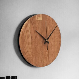 May include: A round wooden wall clock with black hands and a gold center. The clock is made of light brown wood with a natural grain pattern.
