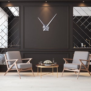 Op de afbeelding: Twee grijze fauteuils met houten frames en een gouden metalen salontafel met een glazen blad. De stoelen staan tegenover elkaar en er staat een vaas met witte bloemen op de tafel. Een grote zilveren klok met lange, dunne wijzers hangt aan de muur achter de tafel.