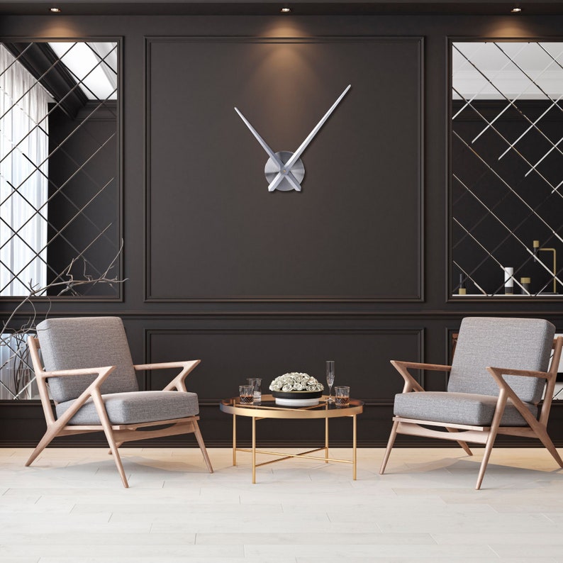 Op de afbeelding: Twee grijze fauteuils met houten frames en een gouden metalen salontafel met een glazen blad. De stoelen staan tegenover elkaar en er staat een vaas met witte bloemen op de tafel. Een grote zilveren klok met lange, dunne wijzers hangt aan de muur achter de tafel.