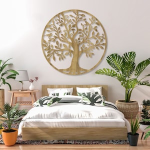 Puede incluir: Una decoración de pared de madera con un diseño circular de árbol de la vida con intrincadas ramas y hojas. El árbol está montado en una pared blanca sobre una cama con ropa de cama blanca y almohadas con estampado verde.