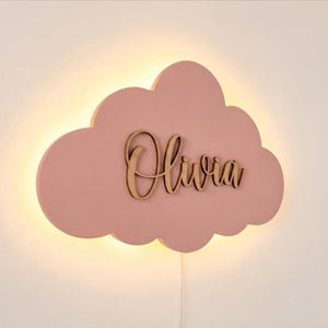 Op de afbeelding: Een wolkvormige wandlamp in een matte oudroze afwerking. In het midden staat de naam Olivia in een donkerbruin, cursief houten lettertype. De lamp verspreidt een zachte, warme gloed van achter de wolkvorm, wat een halo-effect op de muur creëert.