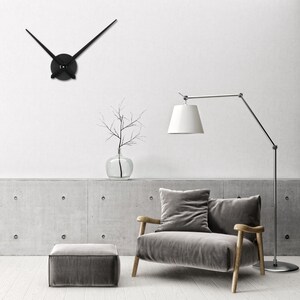 Op de afbeelding: Een zwarte klok met lange, dunne wijzers hangt aan een witte muur. Een grijze fauteuil met een kussen staat voor een betonnen muur. Een staande lamp met een witte kap staat naast de fauteuil. Een kleine grijze poef staat voor de fauteuil.