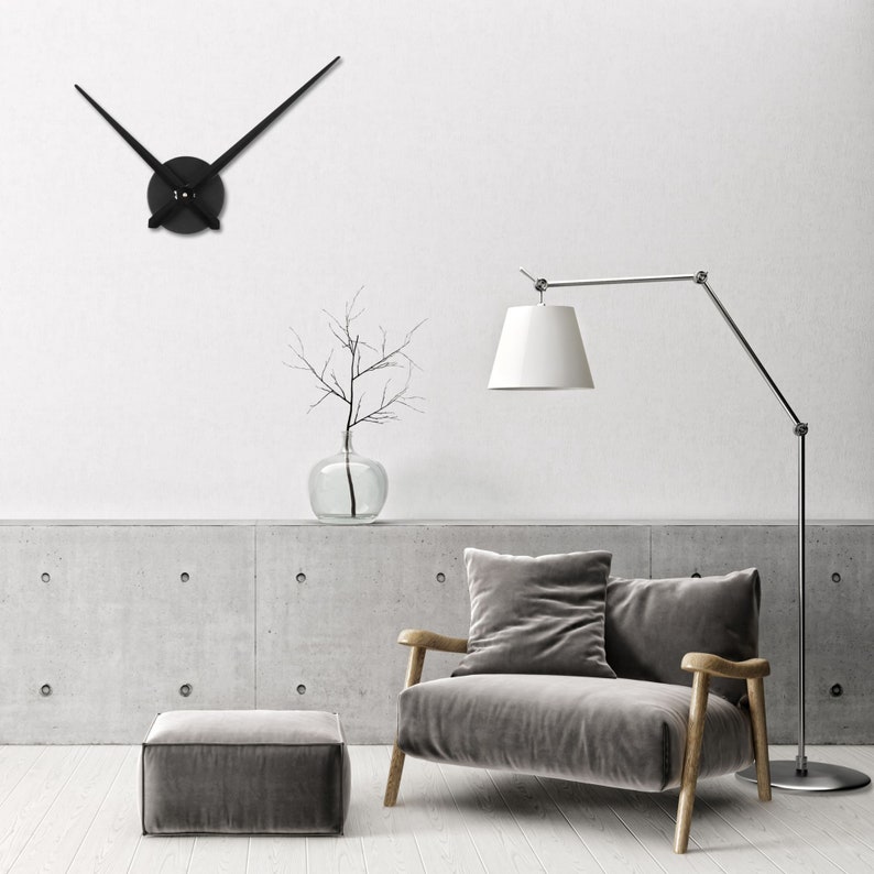 Op de afbeelding: Een zwarte klok met lange, dunne wijzers hangt aan een witte muur. Een grijze fauteuil met een kussen staat voor een betonnen muur. Een staande lamp met een witte kap staat naast de fauteuil. Een kleine grijze poef staat voor de fauteuil.