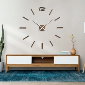 May include: A large, brown wooden clock with white hands and Roman numeral markings on a white wall. The clock is mounted above a wooden entertainment center with white drawers and a shelf. A blue rug is on the floor in front of the entertainment center.