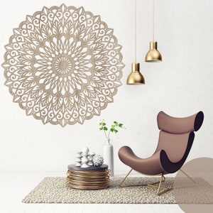 Op de afbeelding: Een groot, beige, cirkelvormig wandkunstwerk met een ingewikkeld, opengewerkt ontwerp dat op een mandala lijkt. De kunst hangt aan een witte muur, aangevuld met gouden hanglampen, een bruine stoel en een kleine tafel.