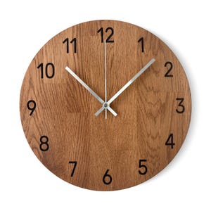 Peut inclure: Horloge murale ronde en bois avec une finition en grain de bois brun clair. L'horloge comporte des chiffres noirs et des aiguilles argentées. Les aiguilles de l'horloge indiquent 12h01.
