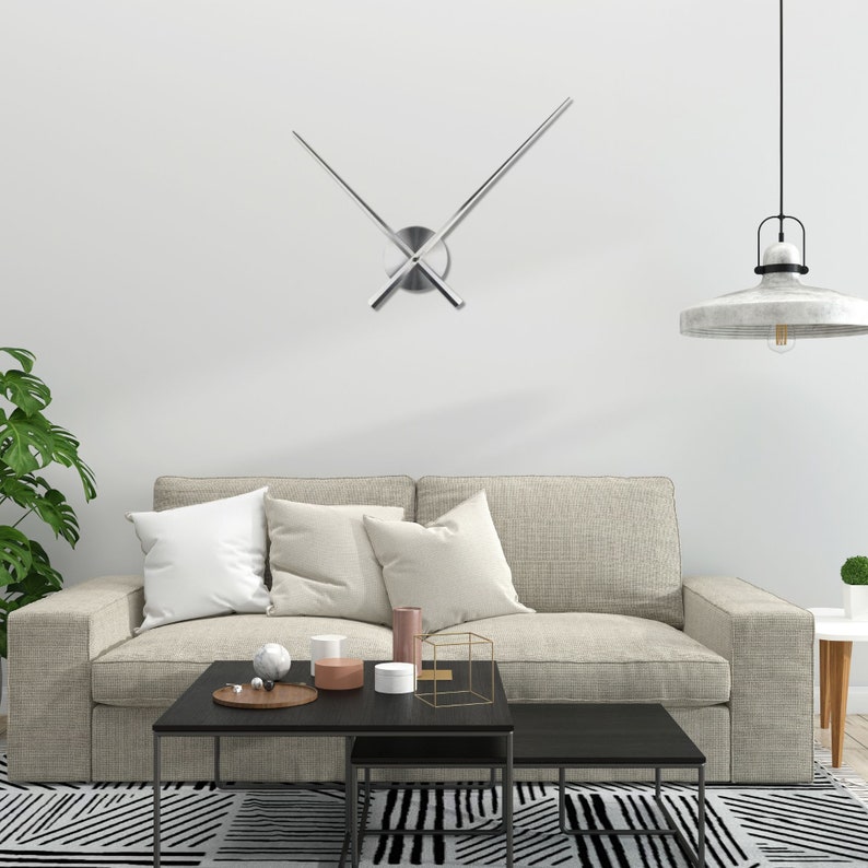 Op de afbeelding: Een moderne woonkamer met een grijze bank, twee zwarte salontafels en een witte muur met een zilveren klok met lange, dunne wijzers.