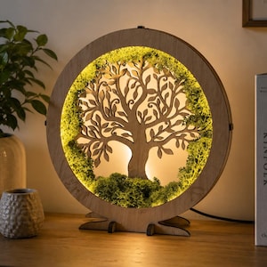 Può includere: Una lampada circolare in legno con una silhouette di albero tagliata al laser al centro. La cornice è rivestita di muschio preservato verde brillante e illuminata da una calda luce LED interna, creando un effetto luminoso dietro i rami.
