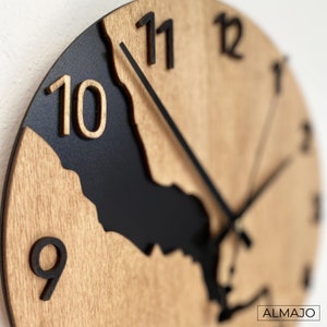 Könnte beinhalten: Eine Holzuhr mit schwarzen Farbakzenten. Das Zifferblatt der Uhr ist halbiert, wobei eine Hälfte schwarz und die andere Hälfte aus Holz besteht. Die Zahlen sind schwarz und die Zeiger sind schwarz. Die Uhr ist rund und hat einen Durchmesser von etwa 30 cm.