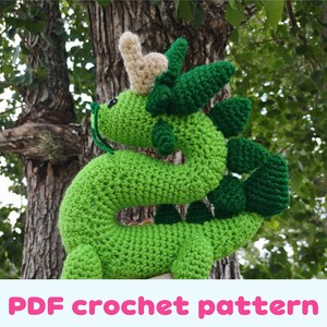 May include: A handmade green crochet dragon with a beige horn and dark green accents. The dragon is posed in front of a tree. The text "PDF crochet pattern" is displayed in pink and white.