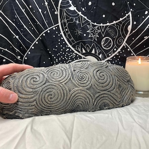 May include: A gray, stone-like sculpture with a spiral pattern, next to a lit candle in a glass. The sculpture is held by a hand. The background features a black and white tapestry with celestial designs. The candle flame glows brightly.