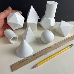 May include: A set of 9 white geometric shapes, including a sphere, cone, cube, cylinder, pyramid, and dodecahedron. The shapes are arranged on a white surface with a wooden ruler and a yellow pencil.