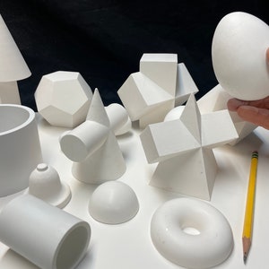 May include: A collection of white geometric shapes, including a cone, a cube, a sphere, a cylinder, a dodecahedron, and a torus. The shapes are arranged on a white surface.