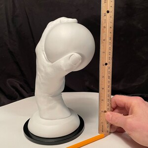 May include: A white sculpture of a hand holding a sphere, mounted on a white base with a black circular bottom. The sculpture is approximately 10 inches tall. A wooden ruler is next to the sculpture.