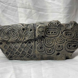 May include: A gray stone sculpture with intricate carved patterns of spirals and circles. The sculpture is shaped like a long, flat boat.