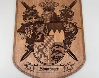 May include: Wooden family crest plaque with a detailed coat of arms design. The crest features a helmet, crossed swords, and a shield with intricate patterns. The name "Stontenger" is displayed on a banner at the bottom. The plaque is made of light brown wood.