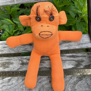 May include: An orange sock monkey doll with button eyes and embroidered details. The handmade toy has a stitched mouth and arms and legs extended. The monkey is sitting on a wooden bench with green foliage in the background.