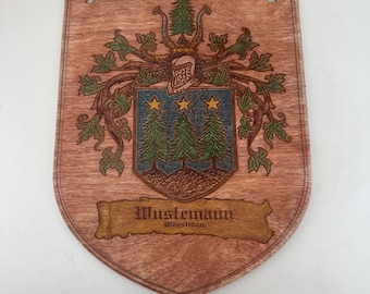 May include: Wooden plaque featuring a family crest design. The crest includes a shield with three stars and two pine trees, a helmet, and a banner with the name "Mustemann". The plaque is brown with a wood grain texture and has two holes at the top for hanging.