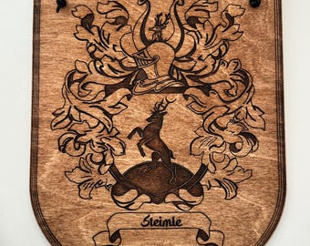 May include: A wooden shield with a coat of arms featuring a deer, a helmet, and floral designs. The shield is stained a dark brown colour and has the name "Steinle" inscribed on a banner.