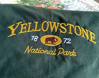 May include: A dark green t-shirt with a yellow embroidered logo that says "Yellowstone National Park" with a buffalo silhouette inside a circle and the years "1872" on either side.
