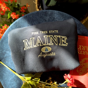 May include: A dark blue sweatshirt with the text "Pine Tree State Maine" and a small sailboat graphic with the text "Augusta" underneath.