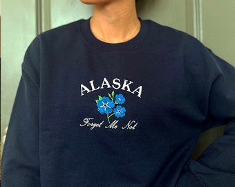 May include: Navy blue sweatshirt with the embroidered word "ALASKA" in white script above a cluster of blue forget-me-not flowers. Below the flowers, the words "Forget Me Not" are embroidered in a delicate script.