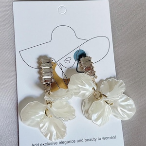 May include: A pair of drop earrings on a white display card featuring a line drawing of a person wearing a hat. Each earring has a rectangular clear crystal stud attached to a cluster of white, translucent, petal-shaped acrylic charms.