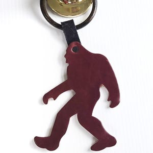 May include: A burgundy leather keychain shaped like a Bigfoot walking. The keychain is attached to a black leather strap and a silver key ring.