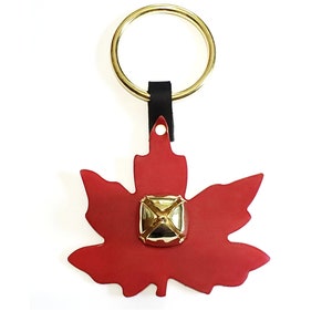 May include: A red maple leaf-shaped door knocker with a gold bell in the center. The knocker is attached to a gold ring and a black leather strap.