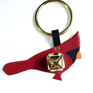 May include: A red cardinal door knocker with a gold bell and a black leather strap. The cardinal is made of leather and has a detailed design.