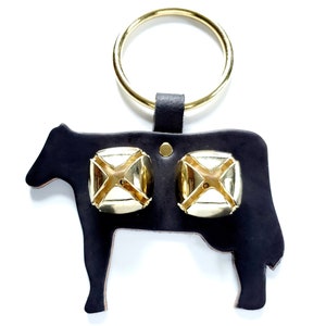 May include: A black leather cow-shaped door knocker with two gold bells. The knocker has a ring attached to the top for hanging.