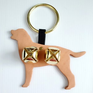 May include: A tan leather dog-shaped ornament with two gold bells attached. A gold ring is connected to the dog by a black strap. The bells are square with a cross design. The ornament is likely for a pet.