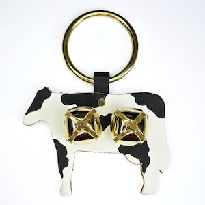 May include: A cow-shaped keychain with a white body and black spots. It features two gold-colored bells and a gold ring for attaching keys. The keychain is made of leather.