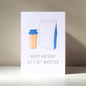 Puede incluir: Una tarjeta blanca con una ilustración azul y naranja de un contenedor de proteína en polvo y una coctelera. El texto en la tarjeta dice "Happy Birthday Let's Get Wheysted".
